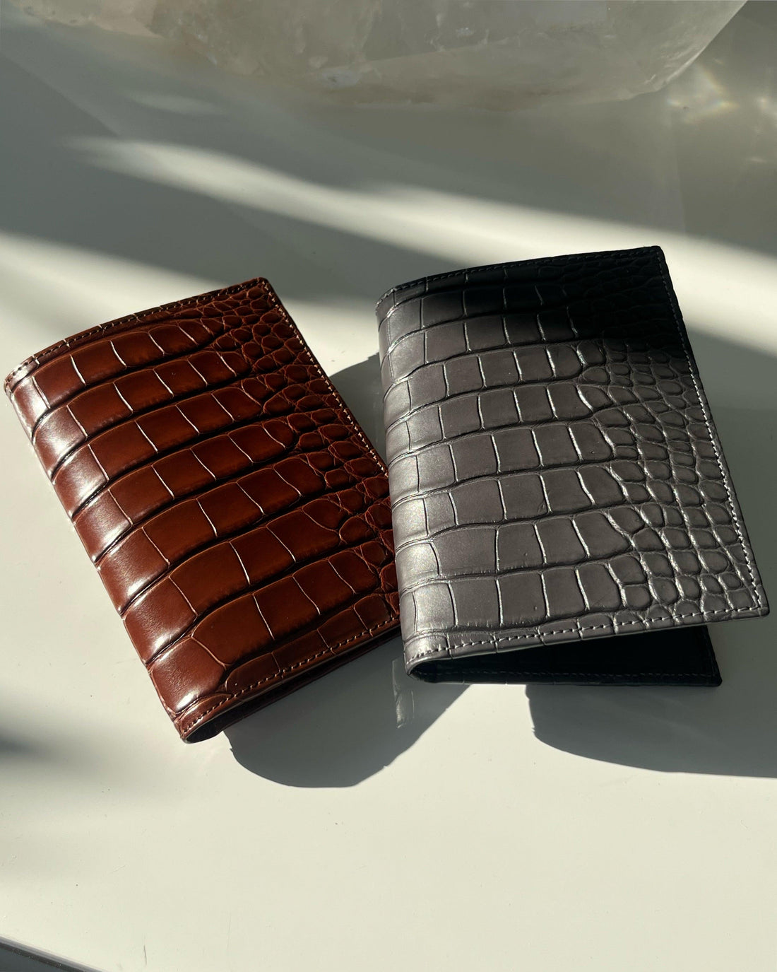 Brown and grey crocodile-embossed leather wallets on a white surface in sunlight.