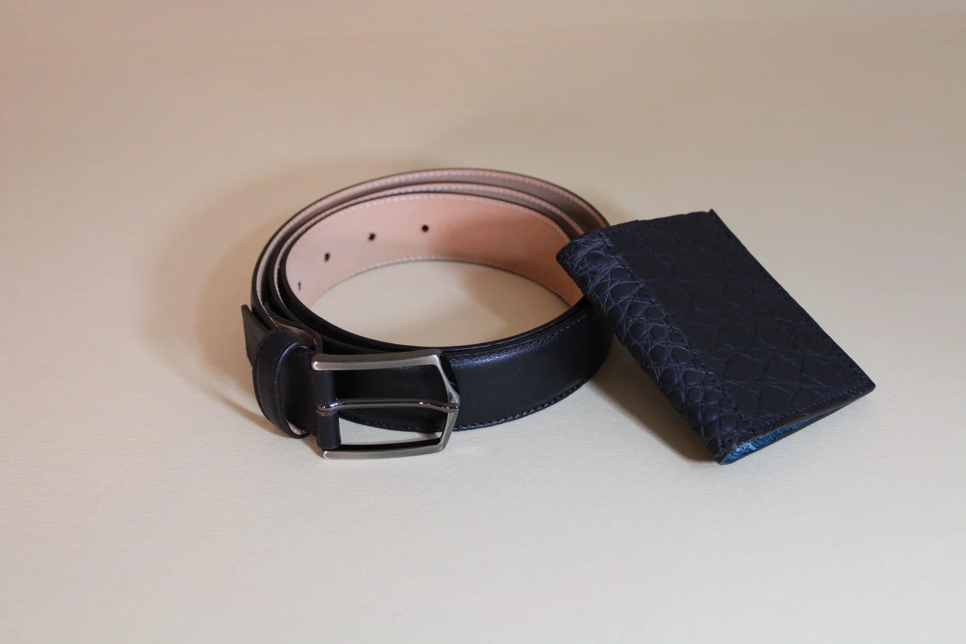 Black leather belt with silver buckle and textured navy leather card holder on beige background