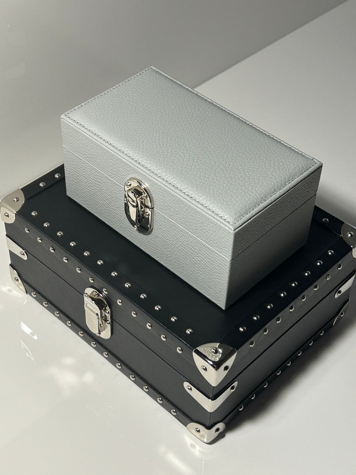 A pale grey leather jewellery case stacked on a studded black leather trunk case