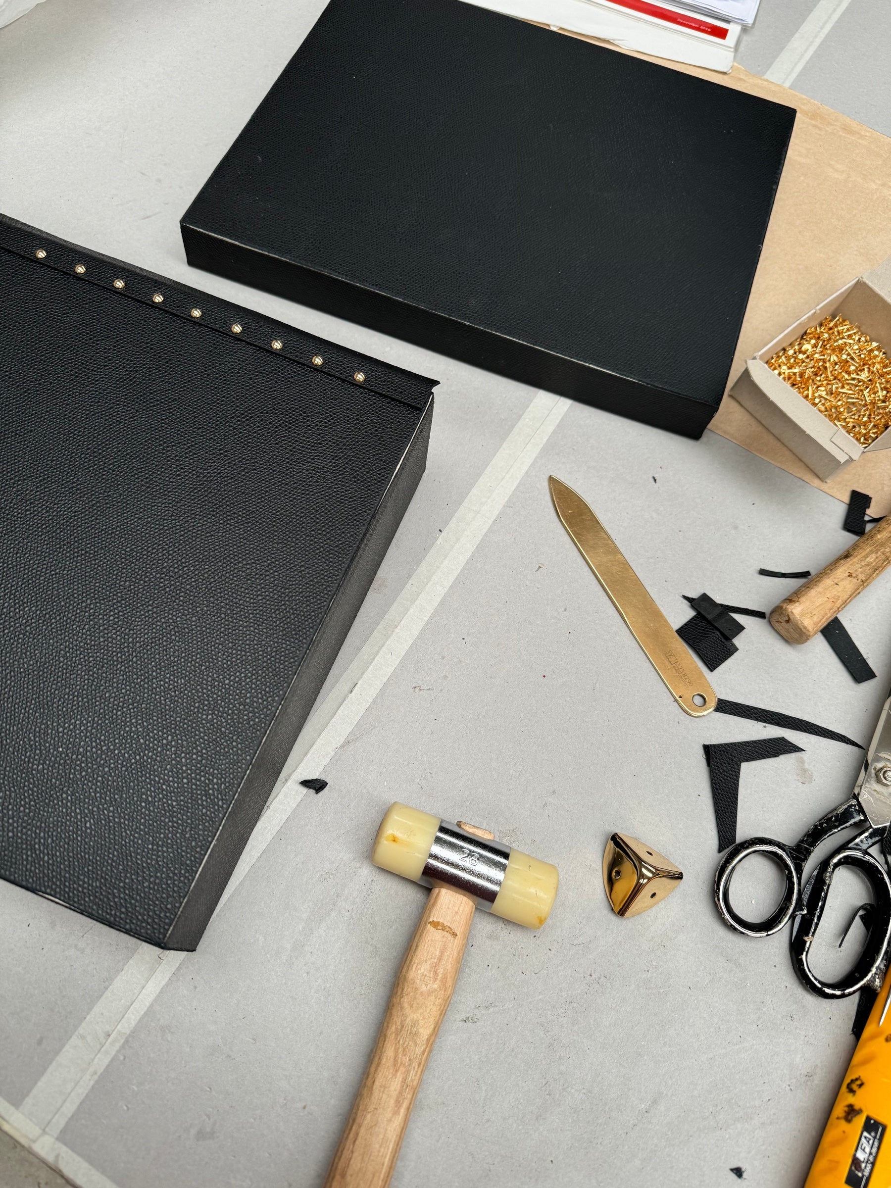Black leather case panels on the atelier bench with brass studs, mallet, shears and a branded brass folder