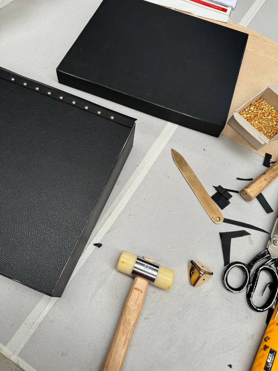 Black leather case panels on the atelier bench with brass studs, mallet, shears and a branded brass folder