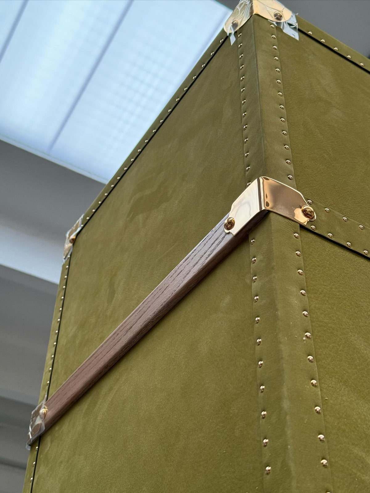 Close-up of an olive-green trunk's stitched corner, brass hardware and handle in a workshop with a skylight